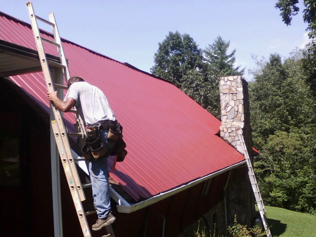 Roofer on a ladder beside a red metal roof and stone chimney — Soffit & Fascia Repair by Roofing Brothers in Cedartown